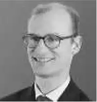 Description de l'image par IA : Homme âgé avec lunettes, sourire, cravate, veste sombre.